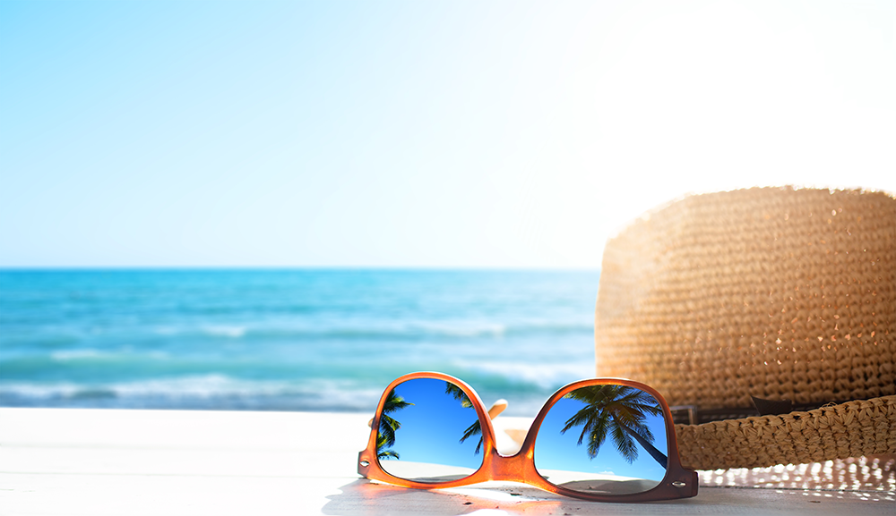 A pair of sunglasses sitting next a sun hat on beach sand, ocean in the background